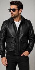 A man wearing a black leather bomber jacket, black sunglasses, a white t-shirt, and black jeans, photographed against a neutral gray studio background.
