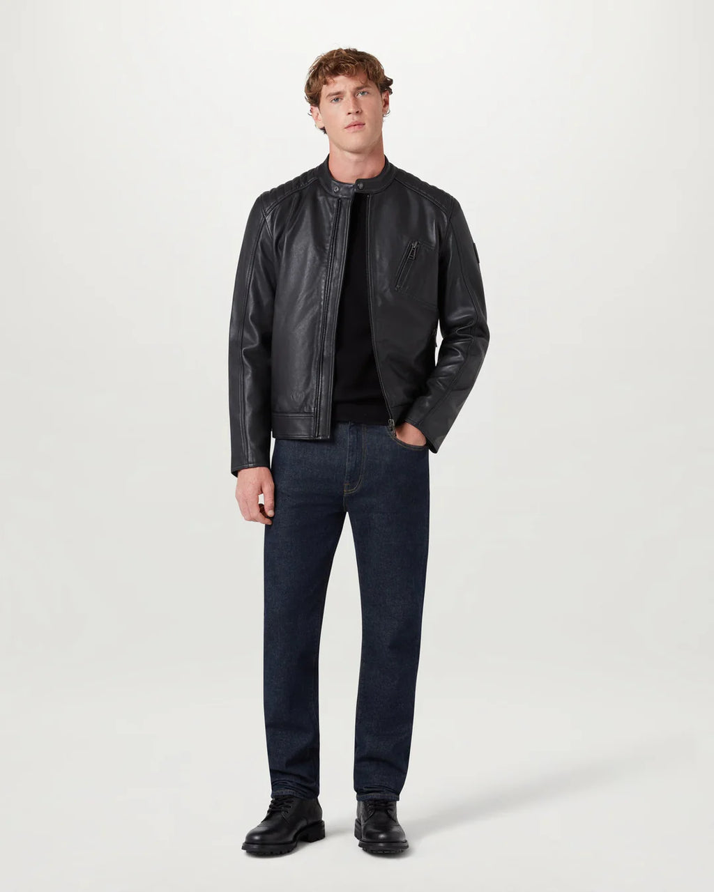 A man modeling a black leather jacket, black t-shirt, dark blue jeans, and black leather shoes against a plain light gray studio background.