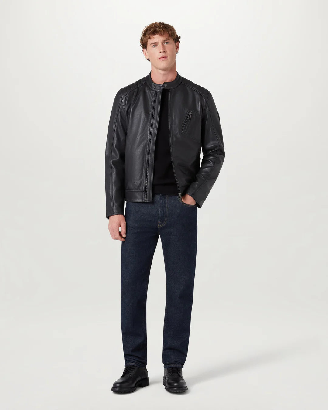 A man modeling a black leather jacket, black t-shirt, dark blue jeans, and black leather shoes against a plain light gray studio background.