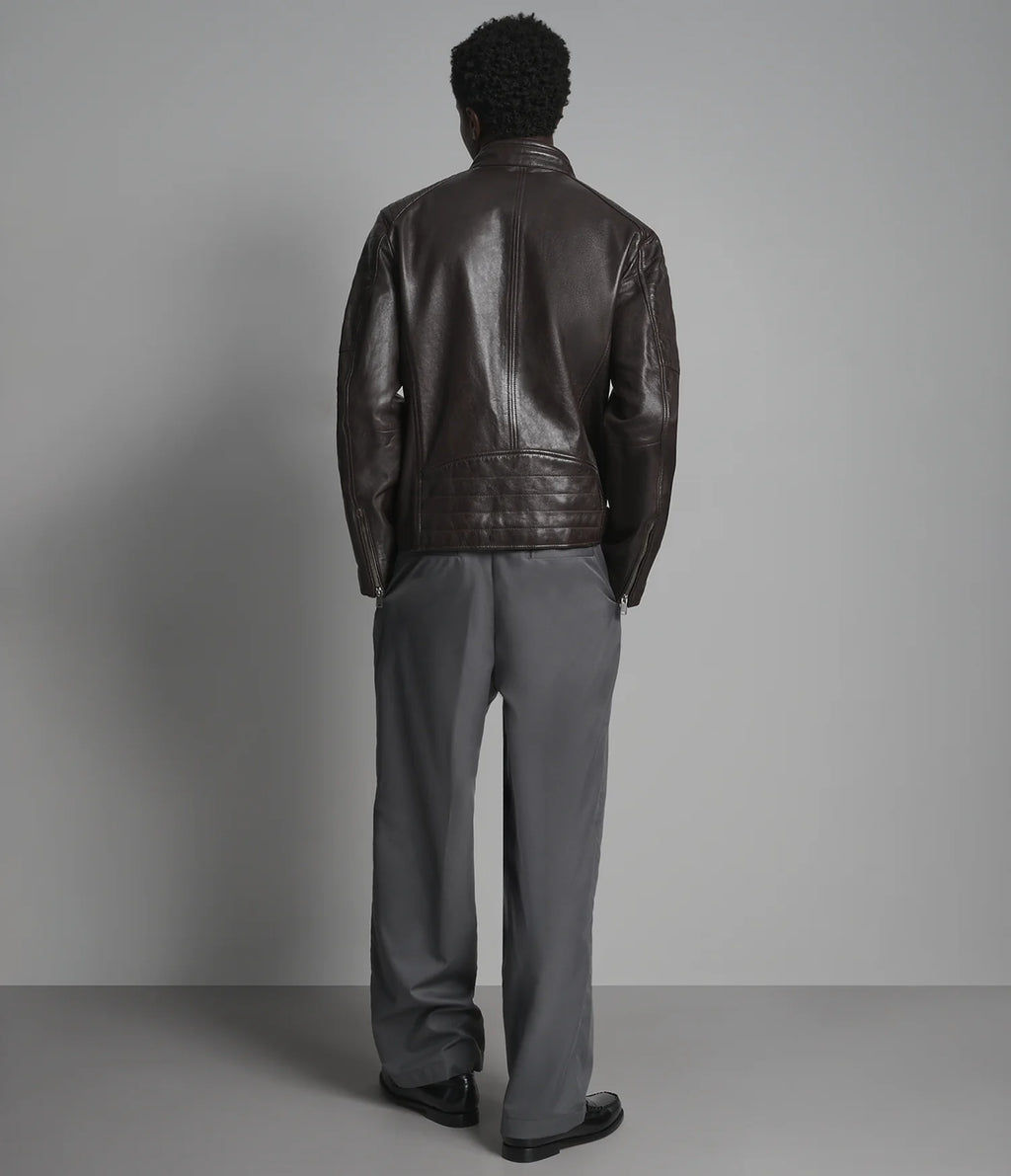 A man viewed from behind wearing a dark brown leather jacket and gray trousers, standing against a neutral gray studio background.