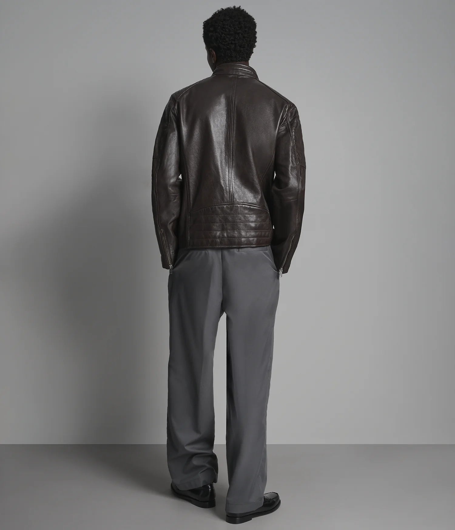 A man viewed from behind wearing a dark brown leather jacket and gray trousers, standing against a neutral gray studio background.