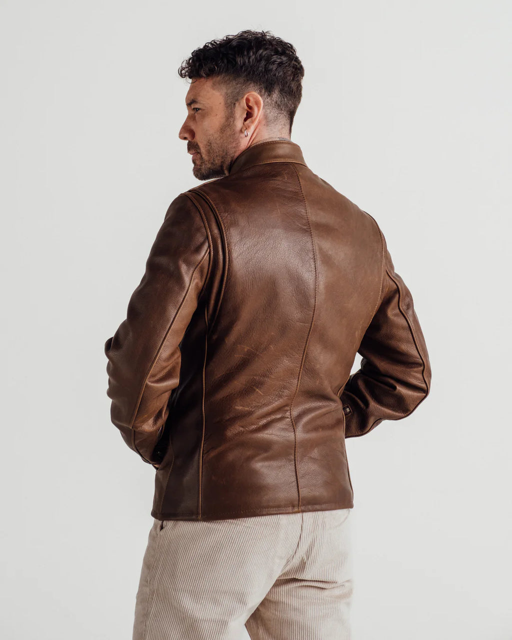 A man wearing a brown leather jacket, shown from the back, paired with light beige trousers, against a plain light gray studio background.