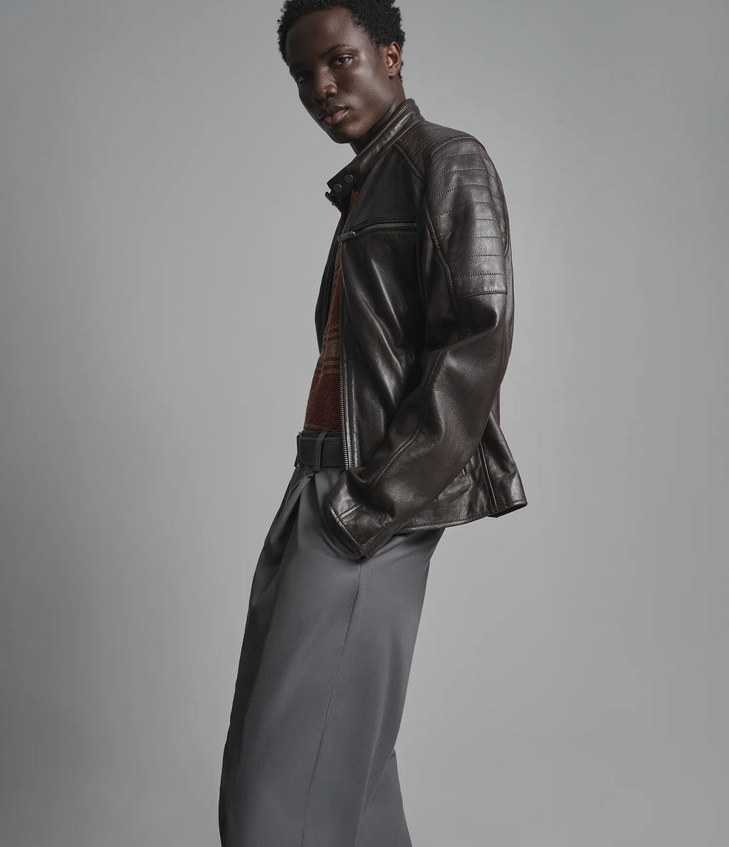 A model wearing a dark brown leather jacket, a rust-colored knit sweater, and gray trousers, photographed against a solid gray studio background.