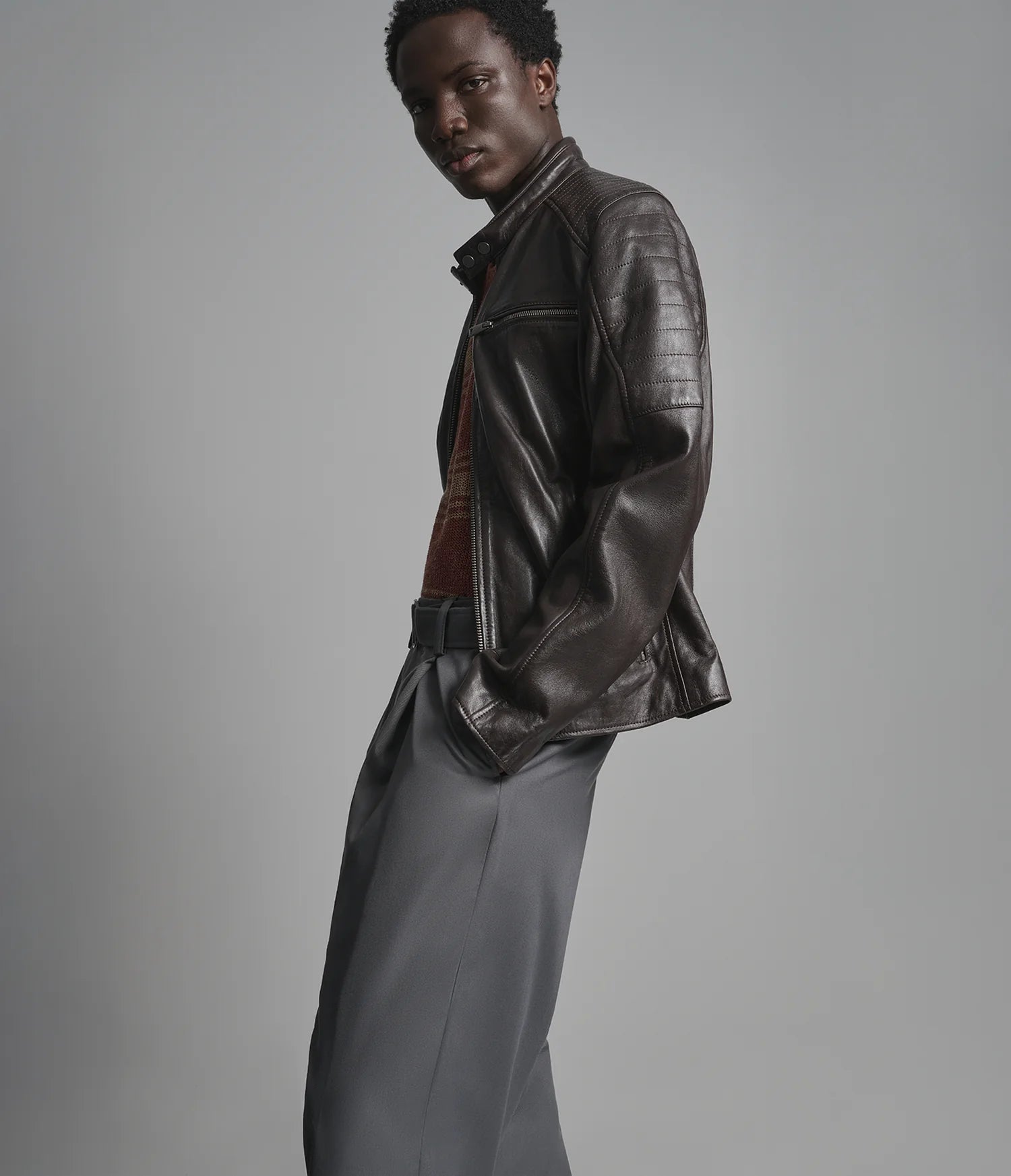 A model wearing a dark brown leather jacket, a rust-colored knit sweater, and gray trousers, photographed against a solid gray studio background.