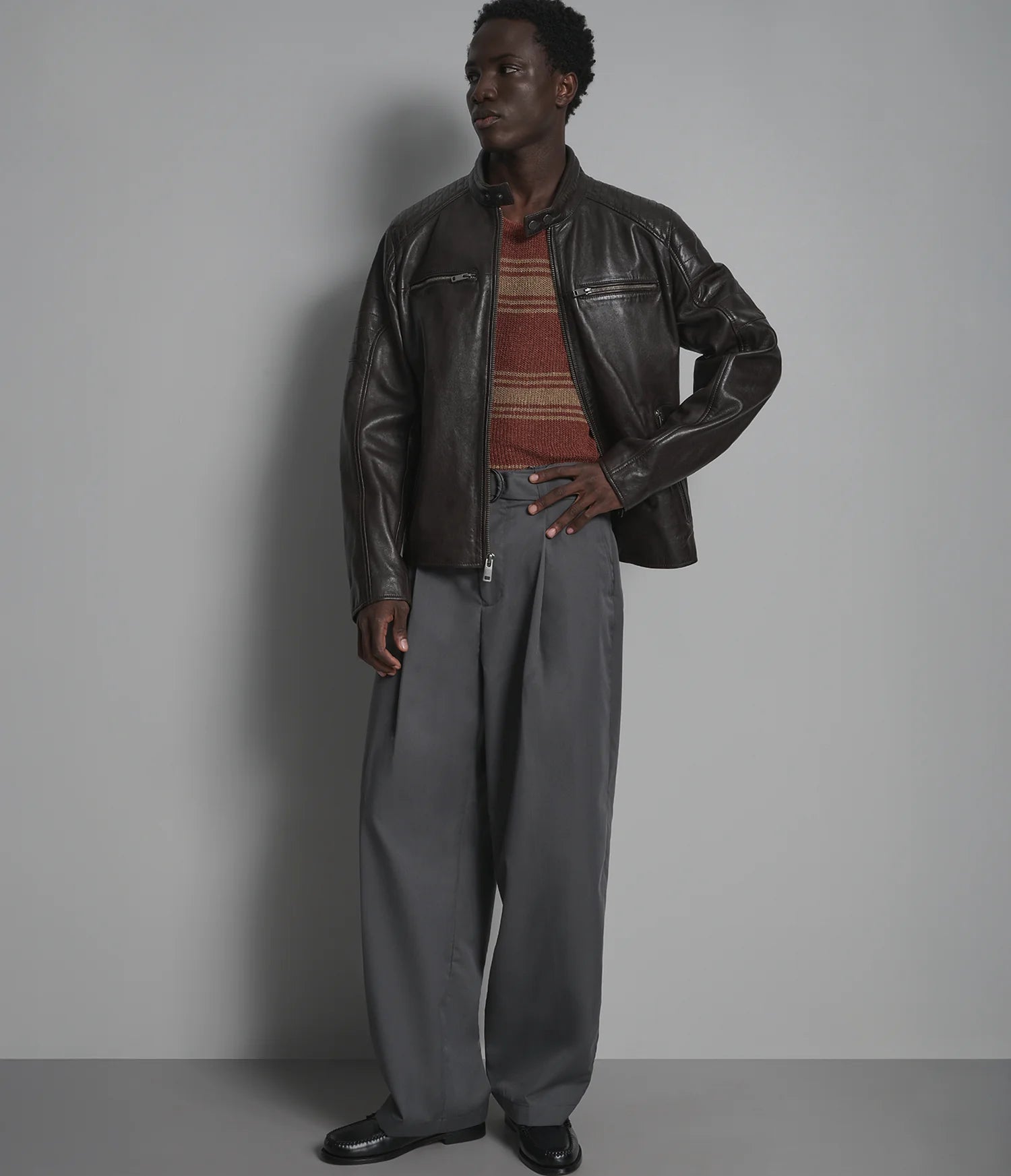 A male model wearing a dark brown leather jacket over a rust-colored striped knit sweater, paired with loose gray trousers and black leather shoes, standing against a plain gray studio background.