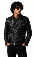 A man wearing a black leather biker jacket, black pants, and sunglasses, standing against a plain white background with his hands in his pockets.