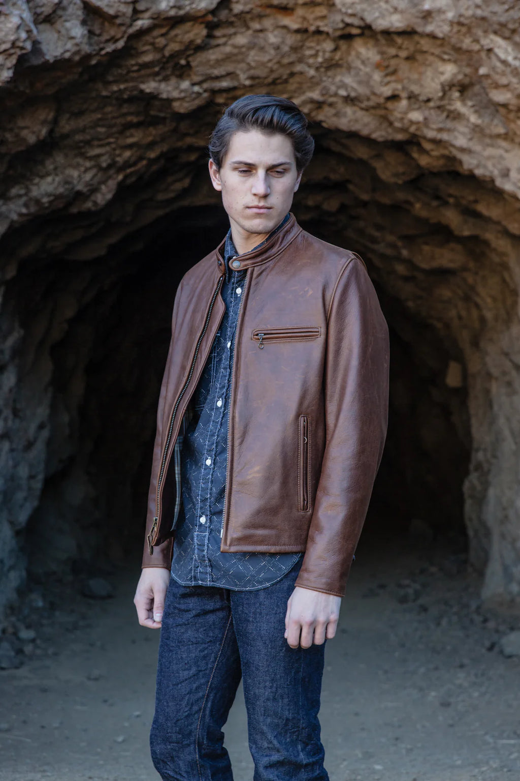 A man wearing a brown leather jacket over a blue button-down shirt and dark jeans, standing in front of a rocky cave opening.