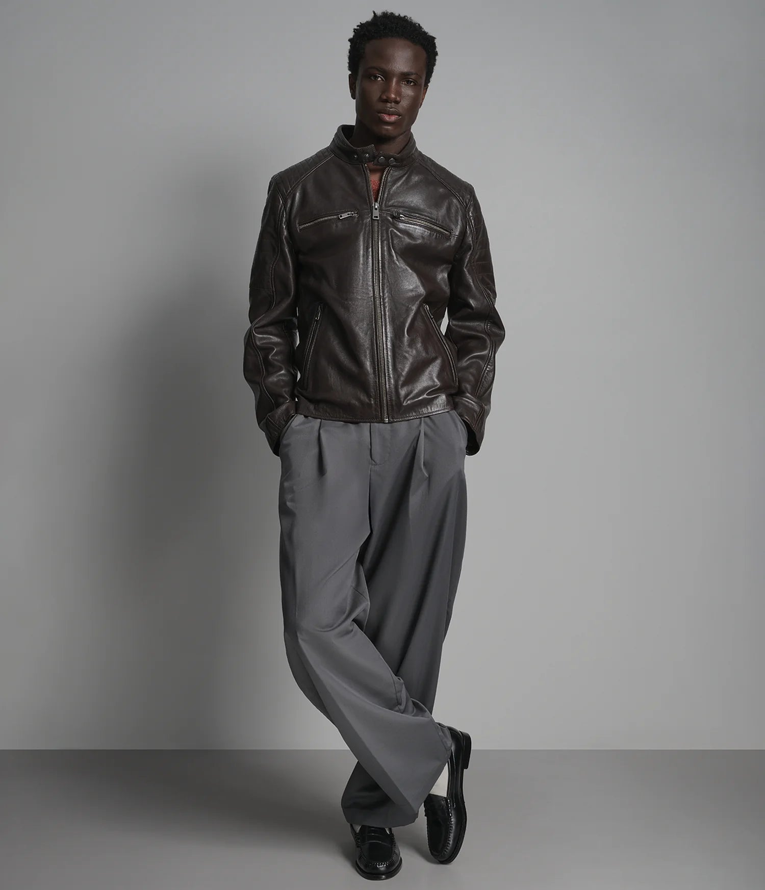 Man wearing a dark leather jacket and gray pants against a plain background