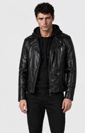 A man wearing a black leather moto jacket over a black hoodie and black pants, standing against a light gray studio background.