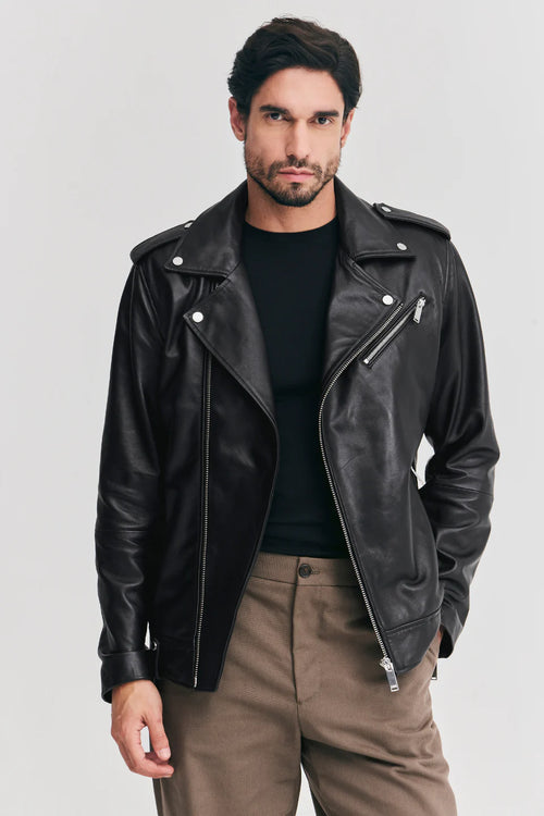 A man wearing a black leather biker jacket over a black t-shirt and brown trousers, standing against a light gray studio background.