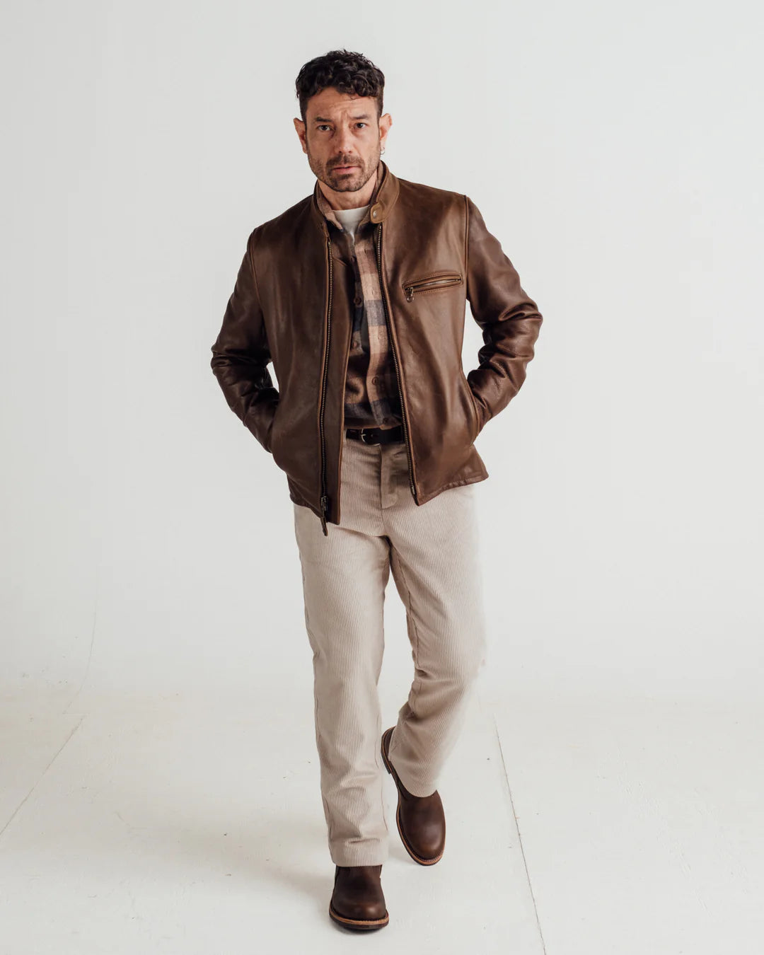 A man modeling a brown leather jacket over a plaid shirt and beige trousers, standing against a plain white studio background.