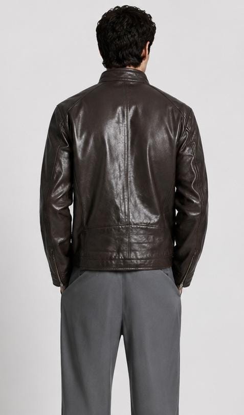 A man viewed from behind wearing a dark brown leather jacket and gray trousers, standing against a light gray studio background.