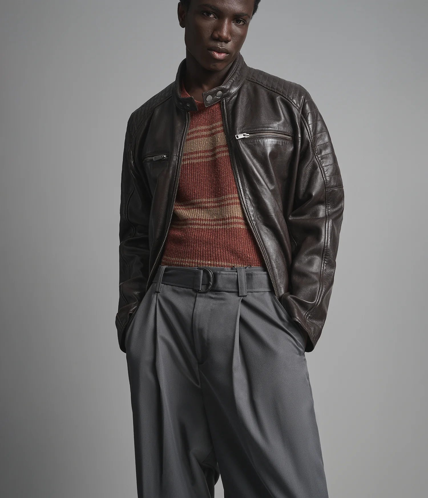A man wearing a dark brown leather moto jacket over a rust-colored striped knit sweater, paired with gray trousers and a black belt, photographed against a neutral gray studio background.