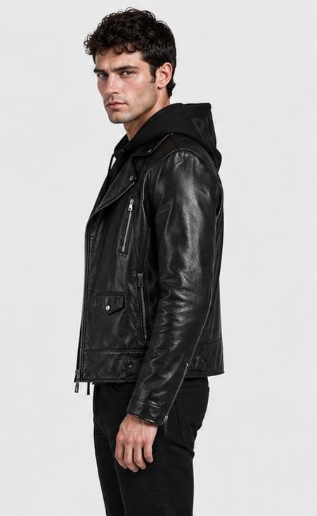 A man wearing a black leather moto jacket with an integrated hood, styled over a black hoodie and black pants, photographed against a plain light gray studio background.